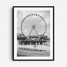 Load image into Gallery viewer, Scheveningen Pier’s ferris wheel with a surfer in front. Black and white North Sea surf photography from The Hague, framed in black wood for a bold and striking wall art piece.
