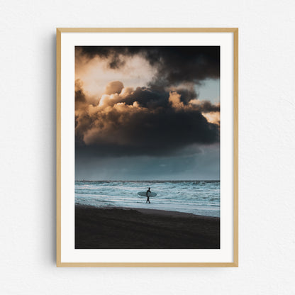 A surfer carries a longboard underneath a heavy sky. Photo print in a natural wooden frame.